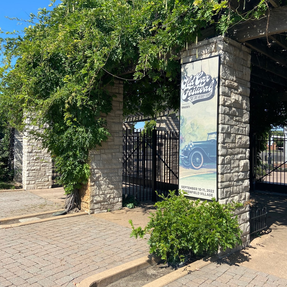 Large stone columns with black iron gates between them and green vines growing down over the tops; one is hung with a banner with text and an image of a car