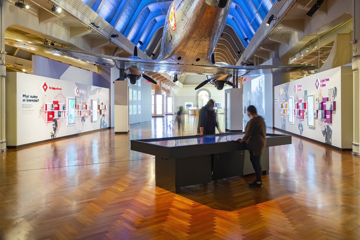 Interior space with two people standing at a large table, graphics and text on the walls, and an airplane above