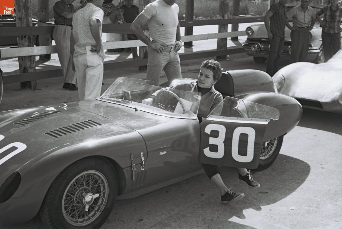 Woman sits with legs out door of small, open race car among other cars and people