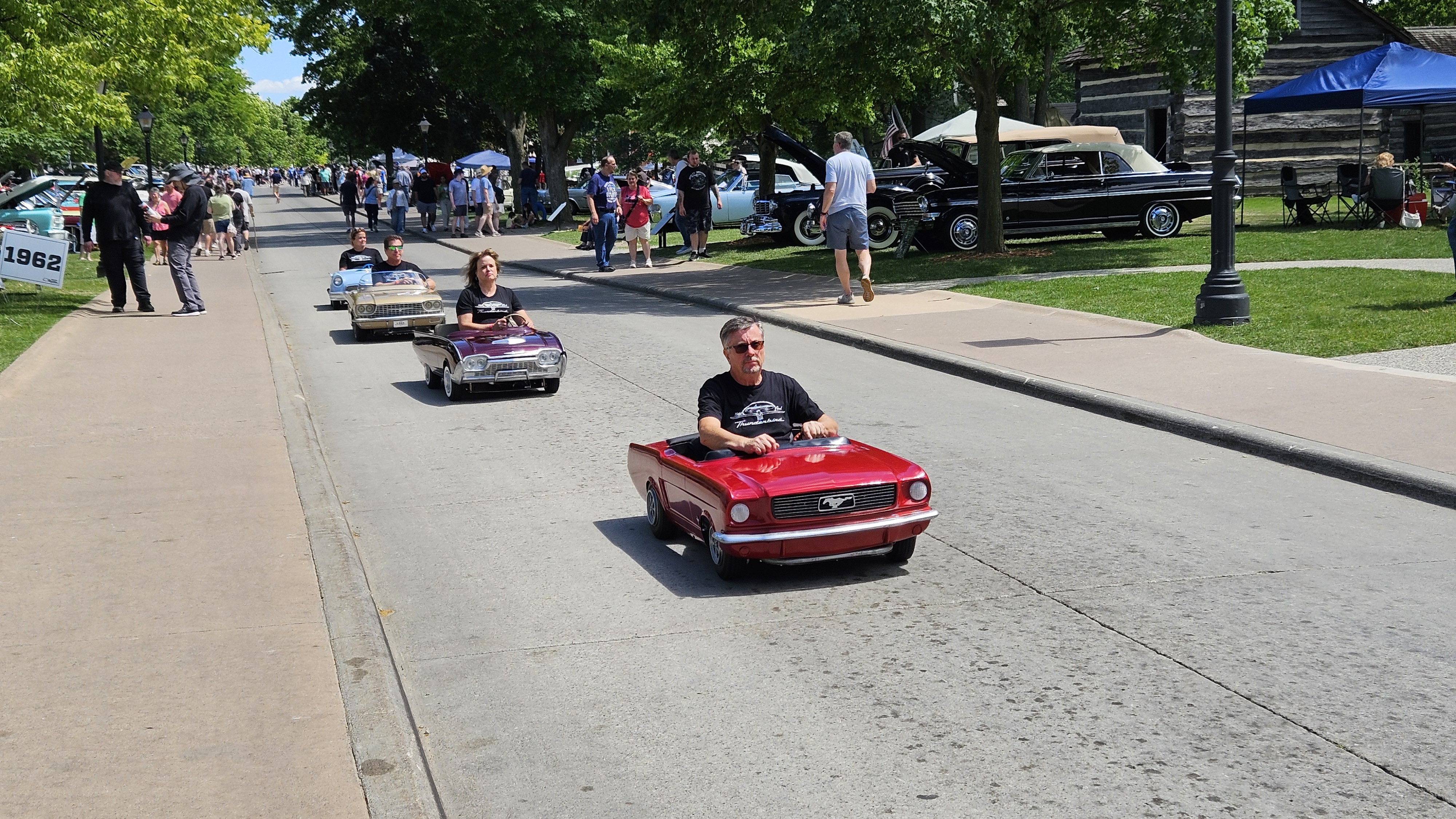 A parade of Thunderbird Jr. kiddie cars led by a Mustang Jr. travels along Maple Lane during Motor Muster 2026, with young drivers enjoying the battery-powered vehicles.