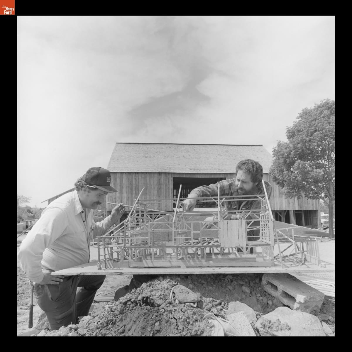 Blake Hayes with a Model of Firestone Barn Used during Its Reconstruction in Greenfield Village, May 1985 Two men look closely at a model of an unfinished building balanced on a pile of dirt with a wooden barn behind them