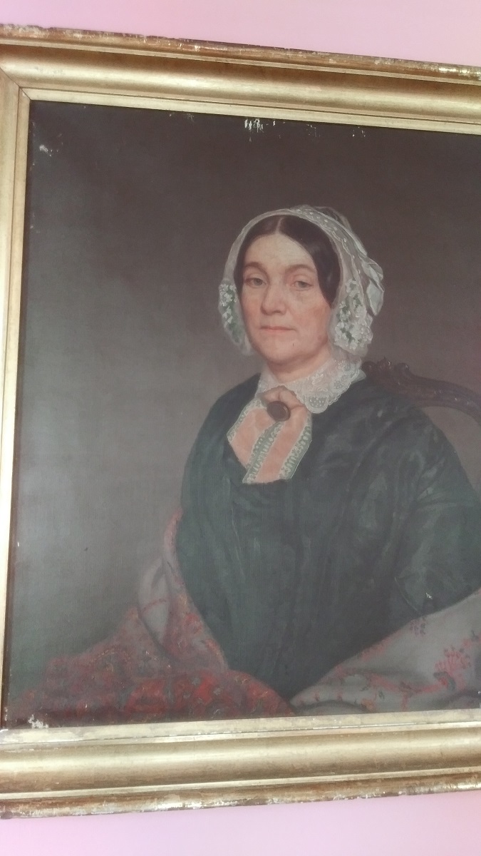 Quick off-center snapshot of painting of woman in black dress, shawl, and white bonnet, showing damage at top of painting