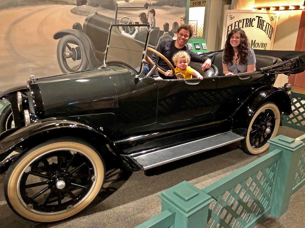 The McAlexander/Flood Family, members of The Henry Ford A man, woman, and child sit in a convertible car with a backdrop and other items in the background