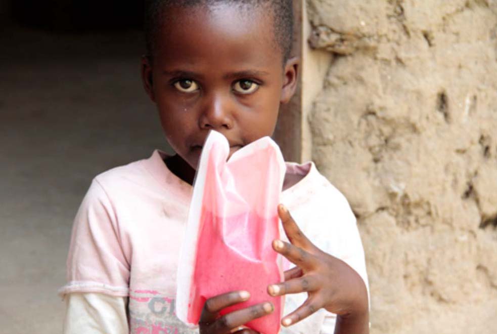 Dark-skinned child in t-shirt holds a packet containing pink liquid to mouth