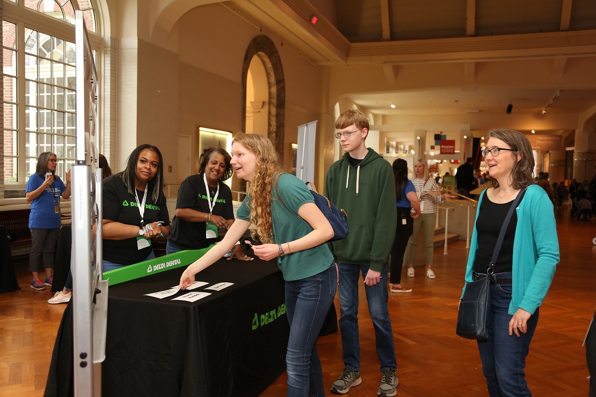 The Delta Dental booth at Invention Convention Michigan Three people look at a sign next to a table draped with a black cloth, behind which are two people in black t-shirts, in a room with other people and displays