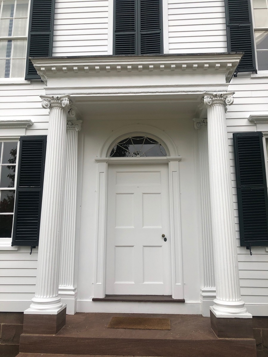 Front door and porch of white building with columns on either side of front door, a small decorative roof over porch, and a decorative half-circle window over the door image001