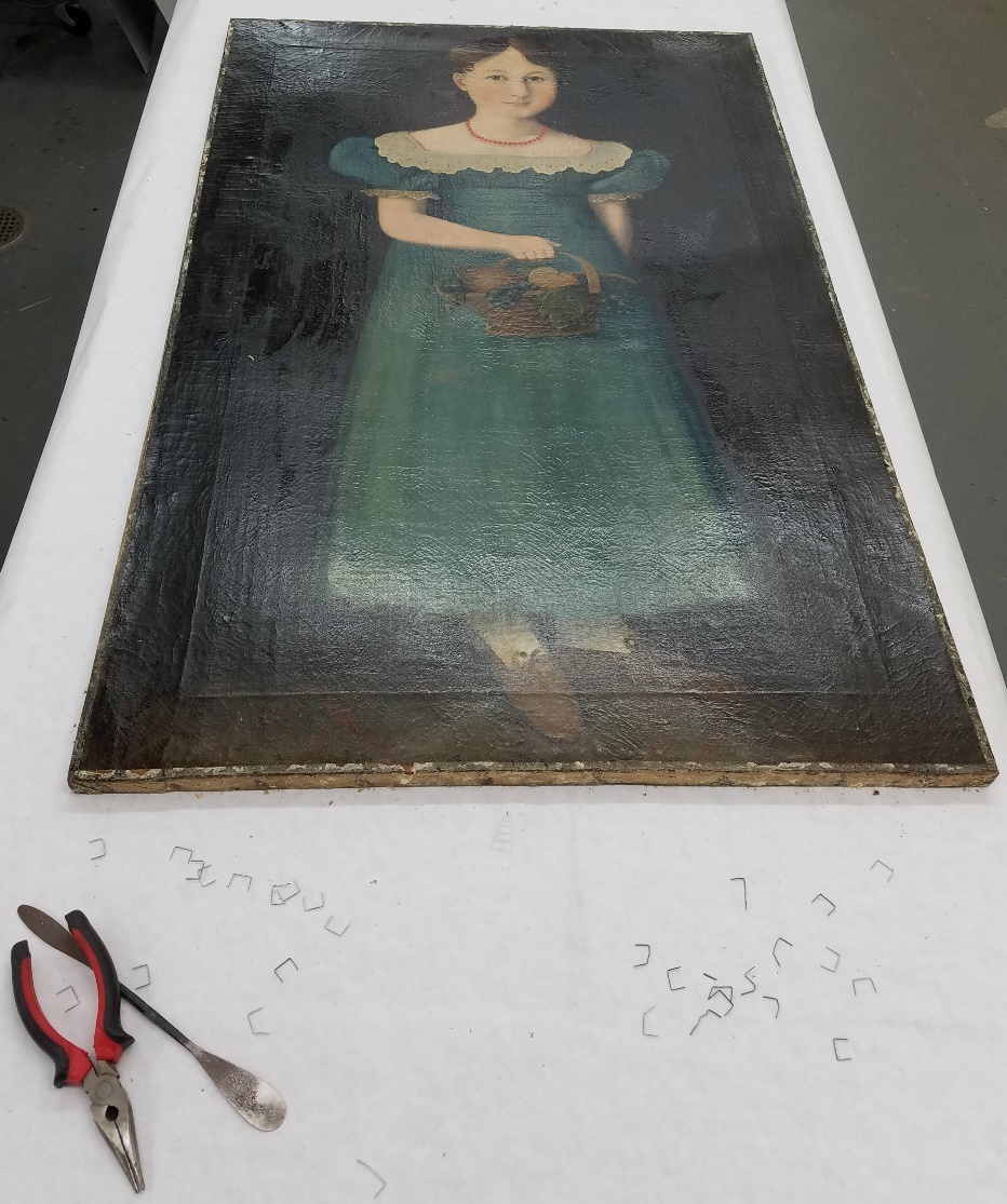 Removing the staples to take the painting off the stretcher Painting of girl in blue dress, laid flat on table with pliers, metal spatula, and staples on table at the end