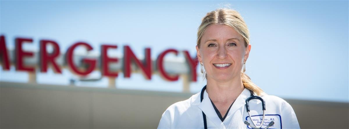 Light-skinned blonde woman in medical coat with stethoscope around neck stands in front of sign reading 