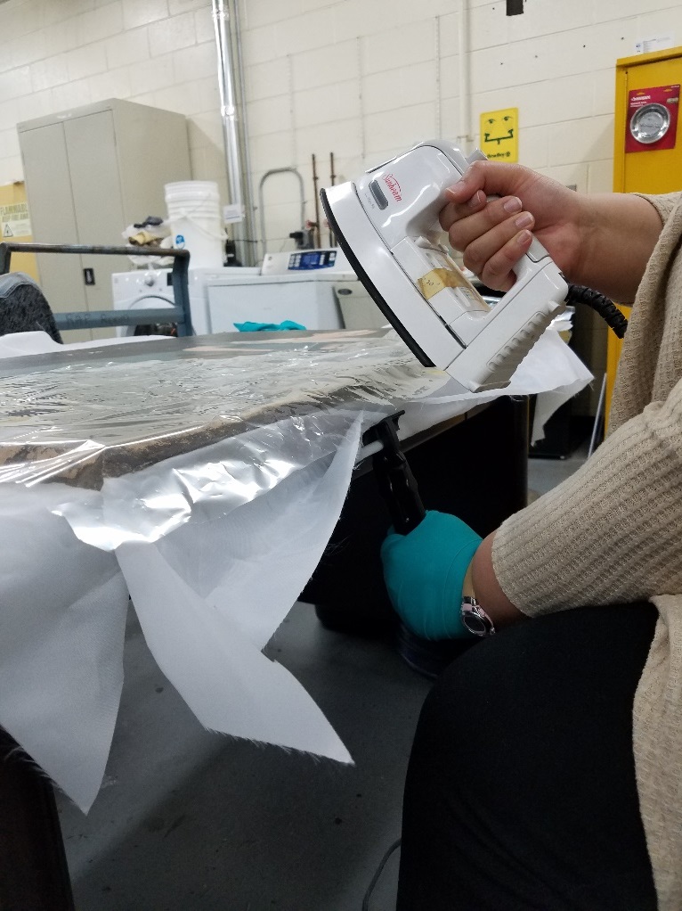 Re-stretching the canvas Person wearing gloves holds an iron to a flat item on a table in a lab