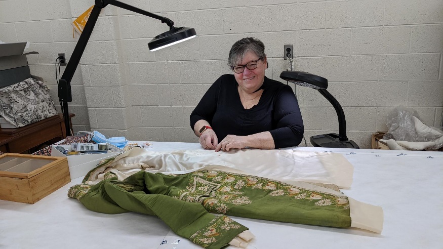Smiling woman in black glasses and black shirt sits at a work table on which is laid out a moss-green jacket with detailed floral embroidery