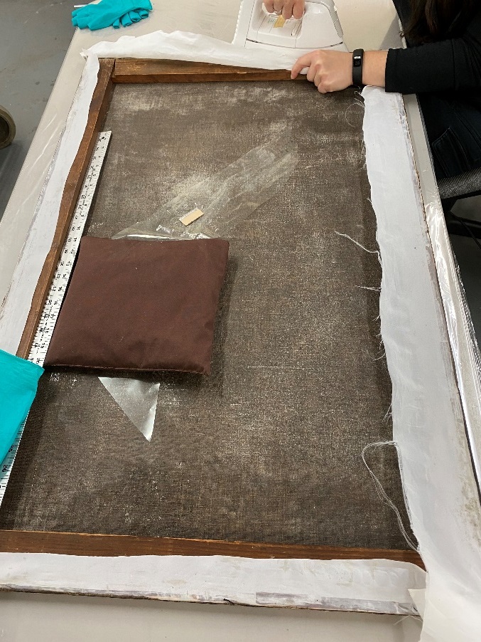 Canvas is re-stretched and extra sail cloth removed A person's hands work at top of painting laid facing down on flat table