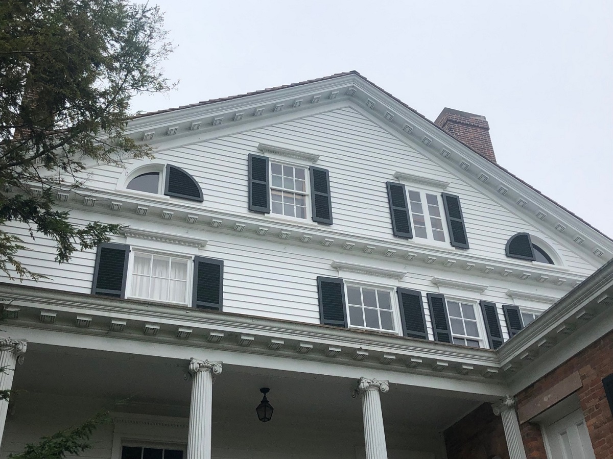 Rear elevation of the Noah Webster Home Peaked gable of a white wooden house with rectangular and quarter-circle-shaped windows; above a porch (?) with columns