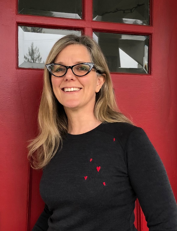 Light-skinned woman with blonde hair and glasses, wearing black sweater with small red hearts, in front of a red door