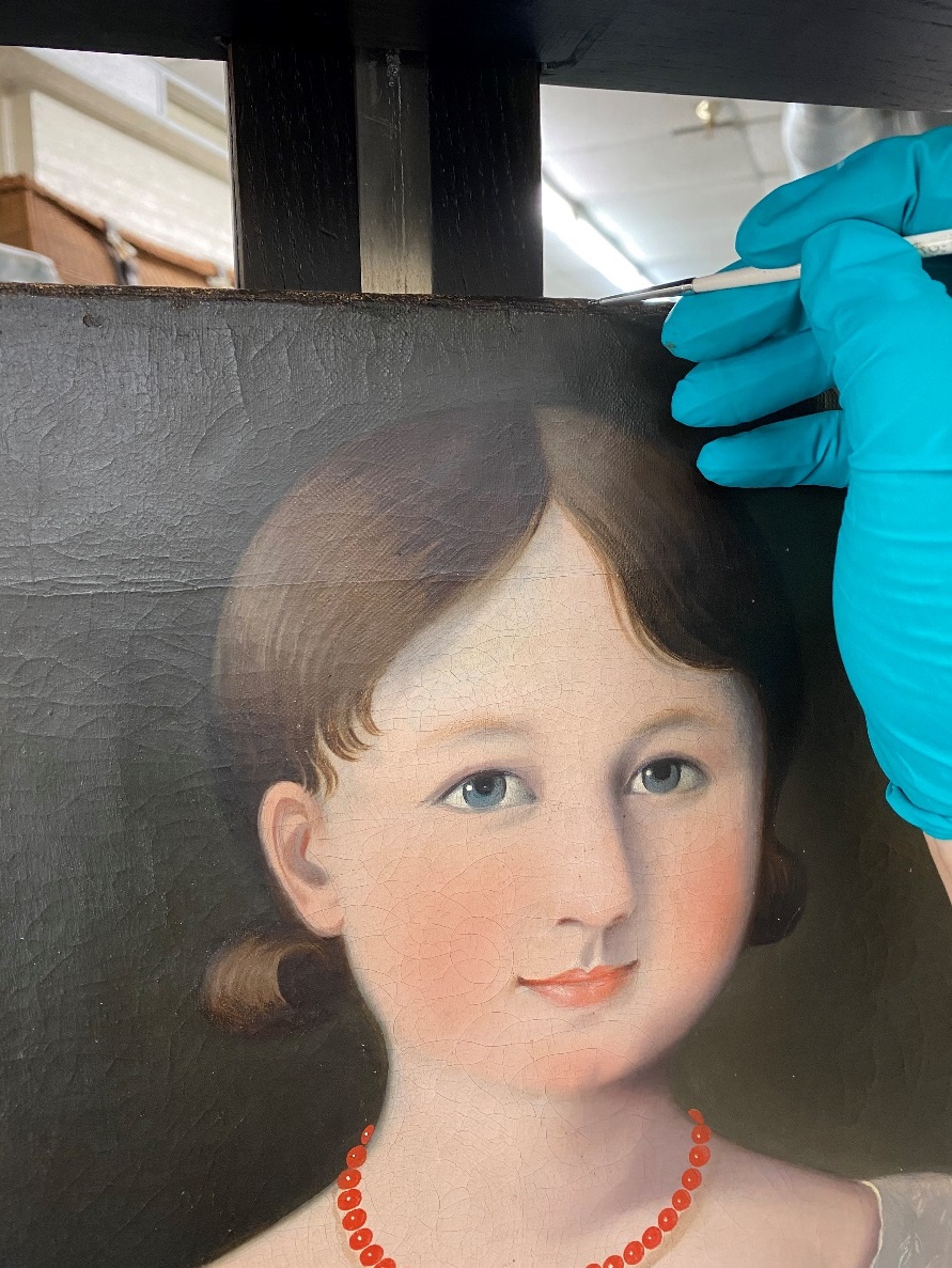 Conservator inpainting Blue-gloved hand holds a paintbrush at the top of a painting of a girl's head