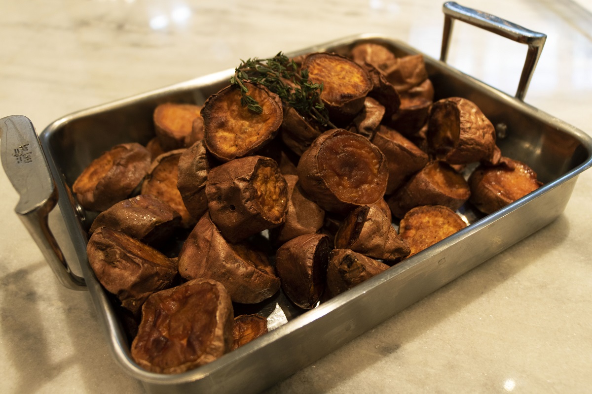 Farmhouse Roasted Sweet Potatoes Silver chafing dish containing large chunks of sweet potatoes topped with herbs, sitting on white and gray marble counter