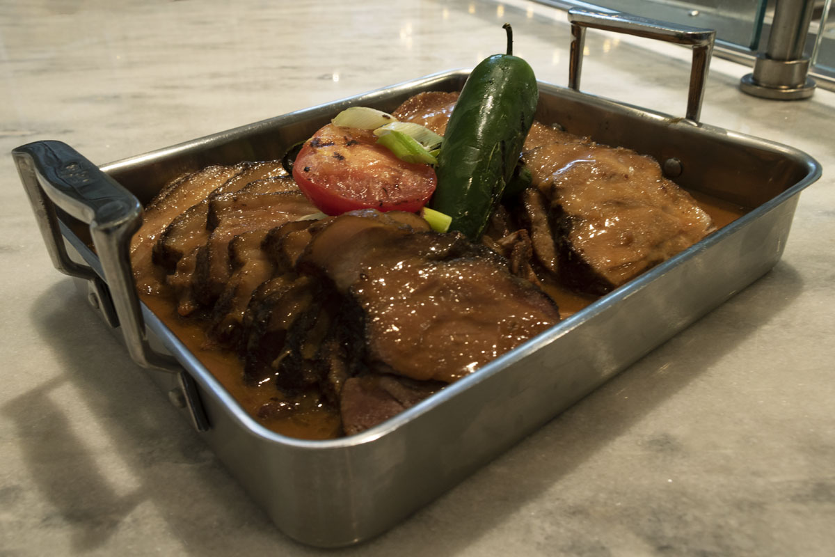 All Natural Pork with Peanut Plum sauce Silver platter filled with meat in a brown sauce, topped with tomato slices and jalapeno peppers; text label behind dish
