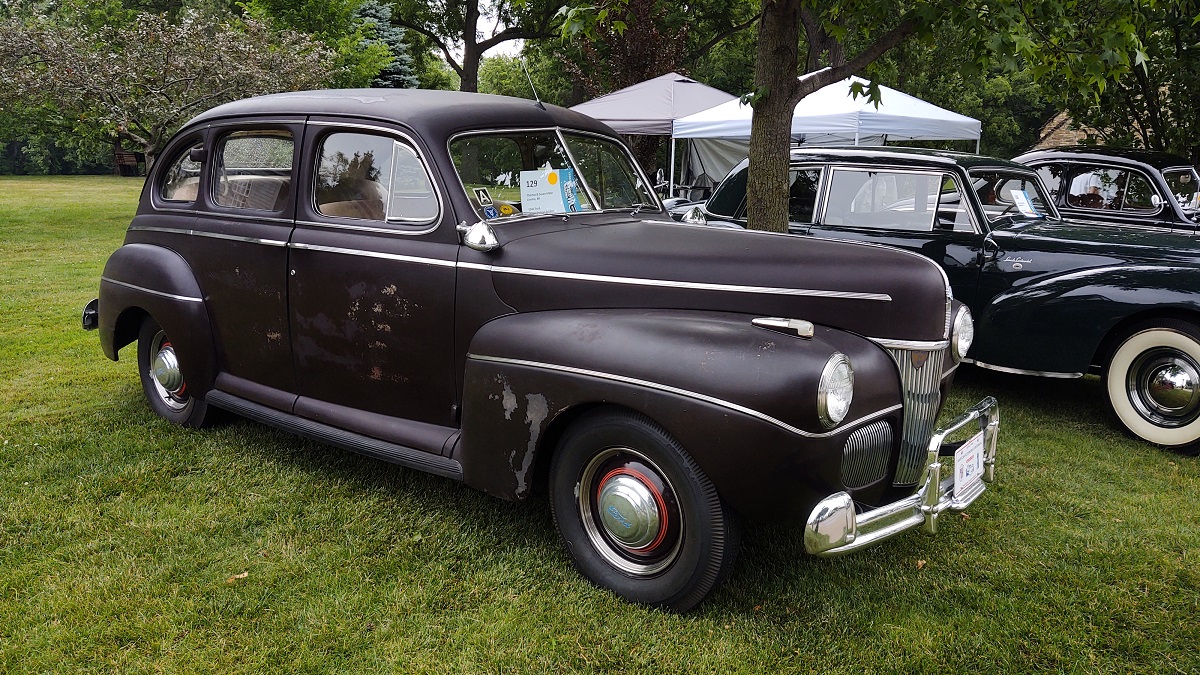 Long dark car with some scuffs in paint, sitting on lawn with other cars and tents nearby