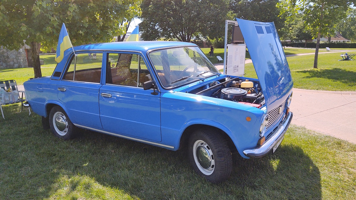 Small, boxy, sky-blue car with hood open on a lawn among trees and walkways