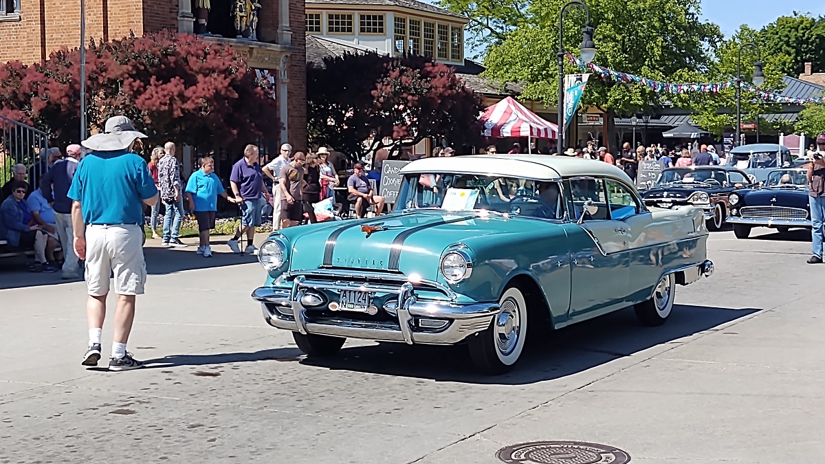 A turquoise and white 1950s vehicle on a road with onlookers and other vehicles nearby