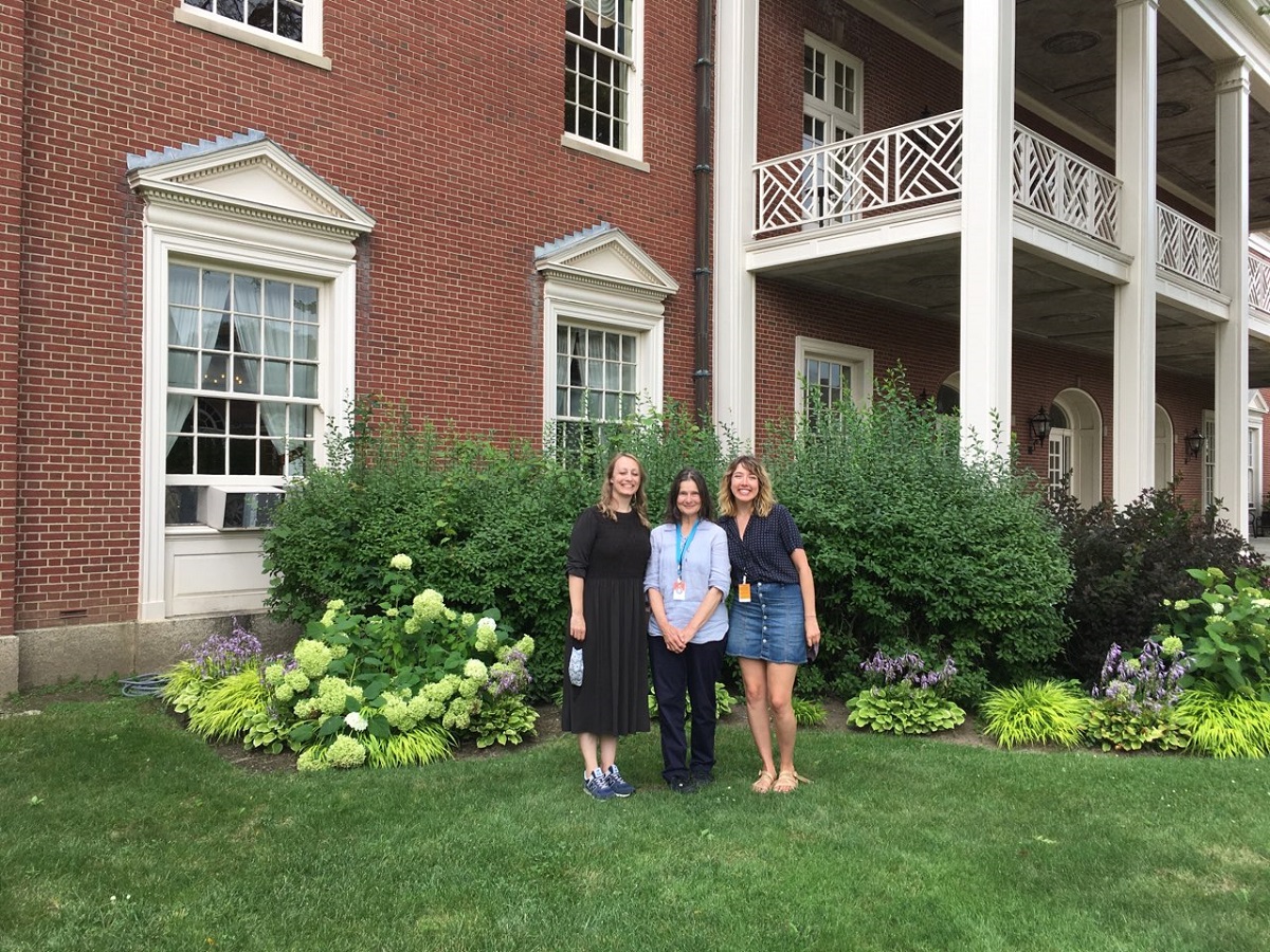 Three women pose in front of flowerbeds and a red brick building with white-pillared porch and balcony