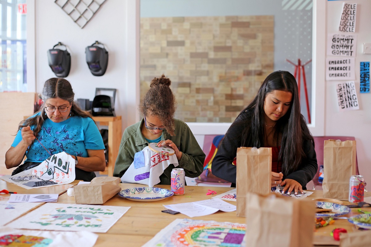 Three people sew at a table full of drawings, plates, soda cans, and paper bags in a large, airy workspace