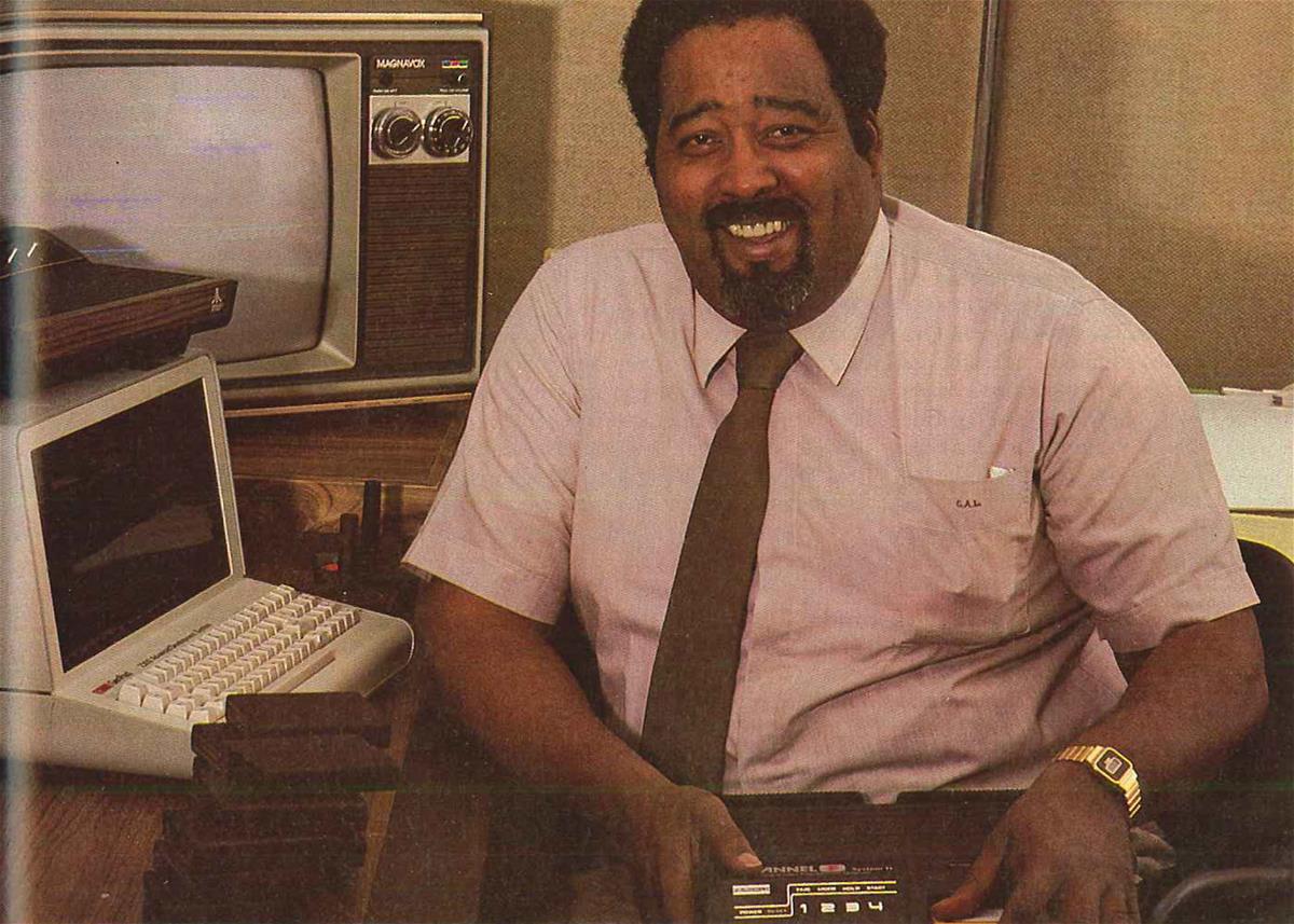 Black man with beard and mustache in shirt and tie sits by a computer and television and smiles at camera