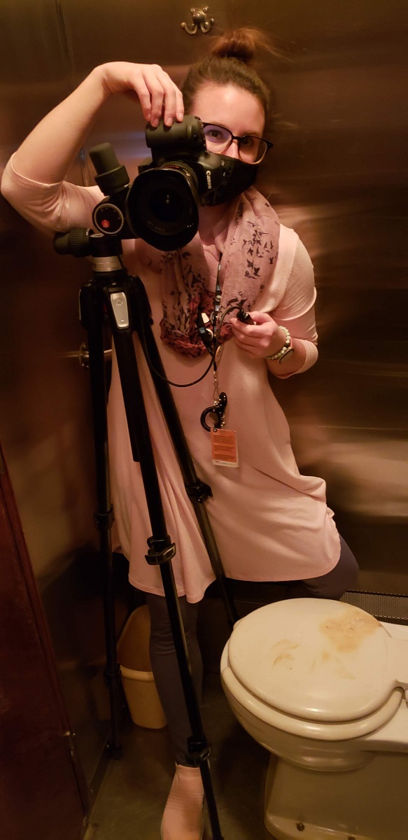 Woman wearing mask behind camera on tripod in a stainless steel room containing a toilet