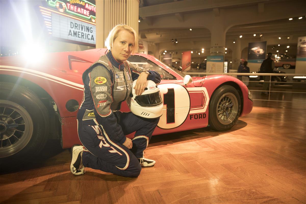 Woman in jumpsuit holding a helmet kneels on wood floor in a building next to low red race car with dramatic backlighting