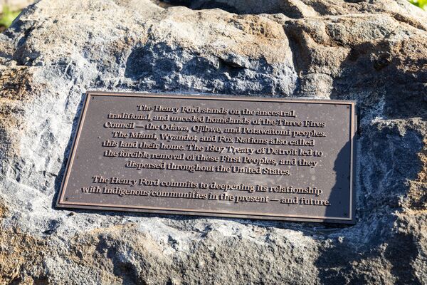 Bronze land acknowledgement plaque mounted on a large natural boulder. The plaque states that The Henry Ford stands on the ancestral, traditional, and unceded homelands of the Three Fires Confederacy—the Odawa, Ojibwe, and Potawatomi peoples—and affirms a commitment to strengthening relationships with Indigenous communities in the present and future.