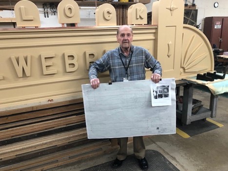 Man wearing plaid shirt holds drawing in front of large sign in workshop space