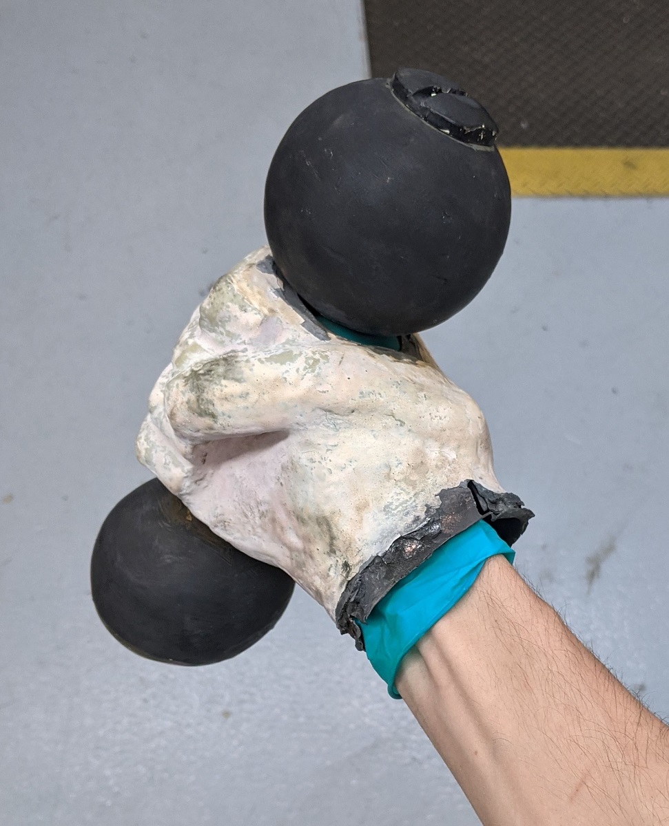Person's wrist wearing blue glove inside white sculpted fist holding a barbell