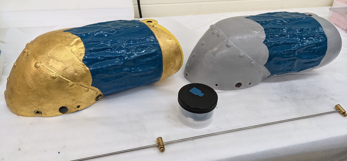 Two identical tubular shapes next to each other, one gold and blue and one gray and blue, with small jar between them