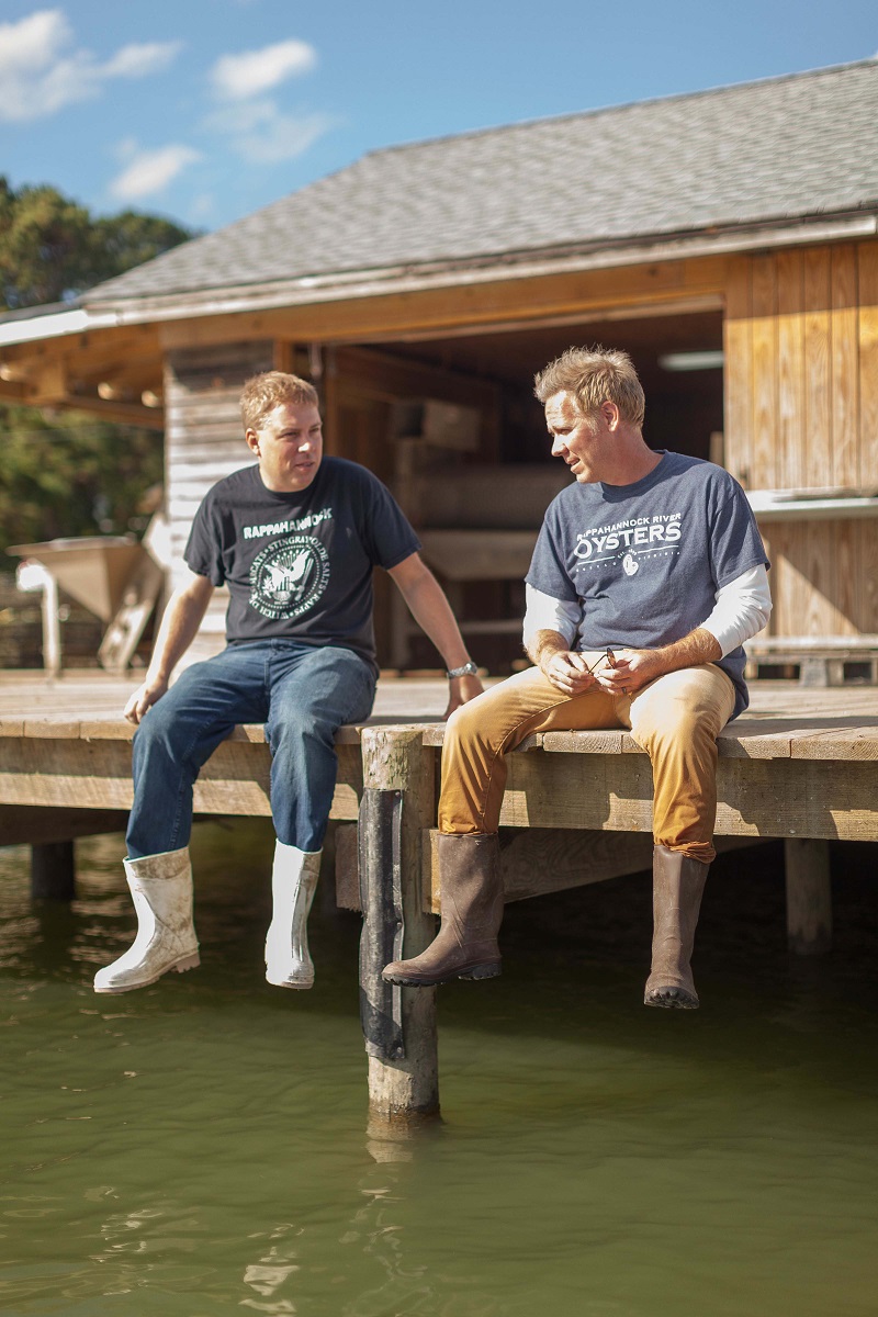 Travis and Ryan Croxton of Rappahannock Oyster Co. Two men wearing boots, khakis/jeans, and t-shirts sit with their legs dangling off a dock with a wooden building in the background