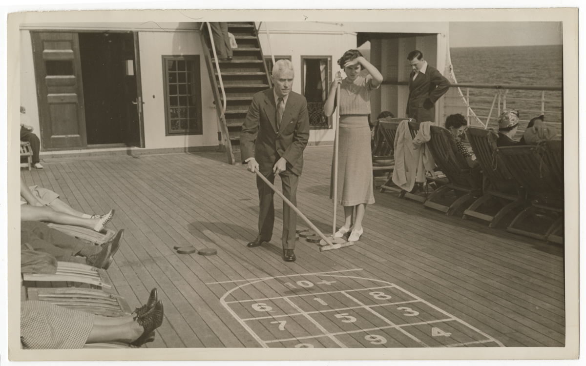 Black and white photo of man in suit and woman in dress playing shuffleboard on a boat with passengers in lounge chairs nearby