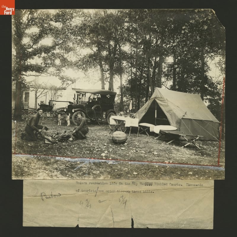 Black-and-white photo of campsite with tent, people by a cooking fire, a Model T, two goats, and two dogs