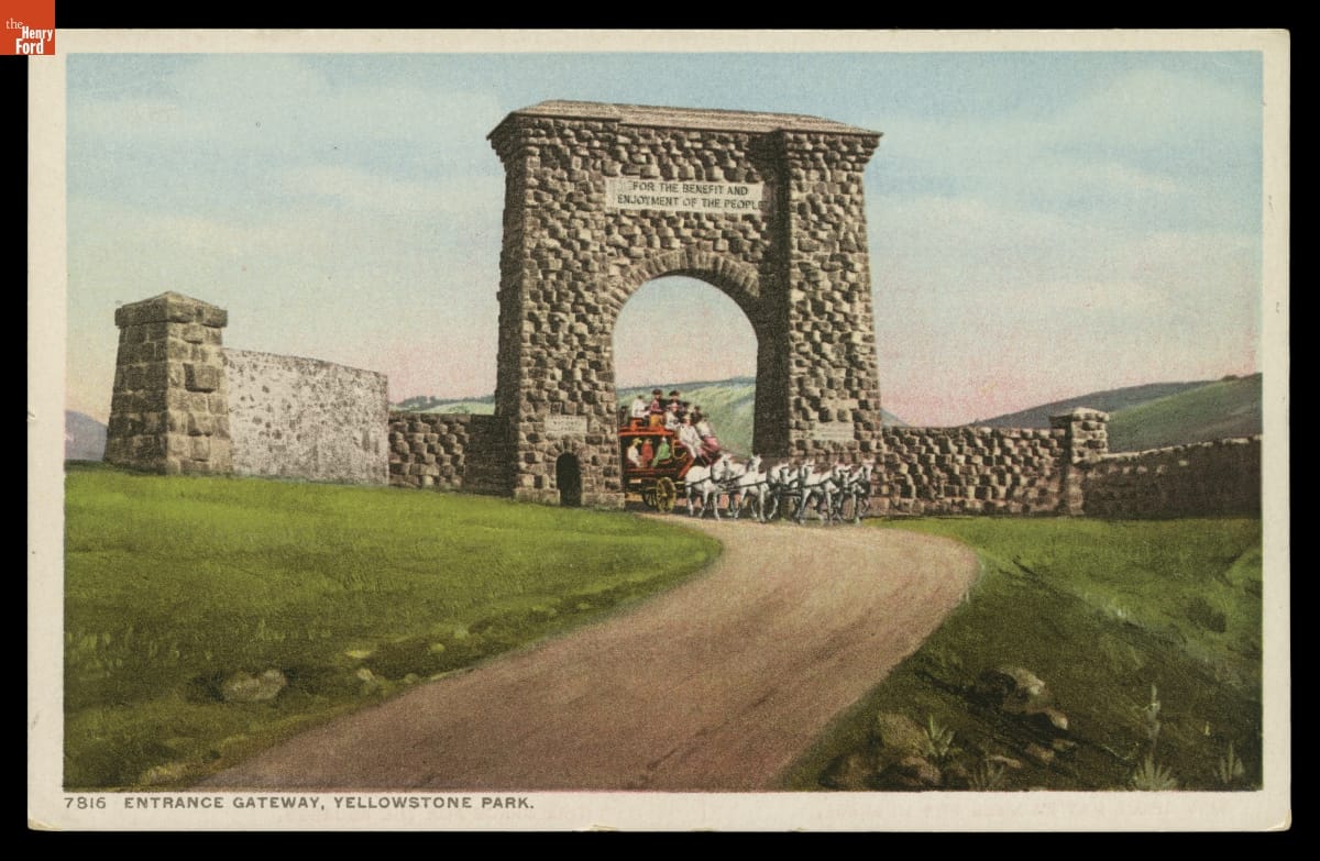 Postcard showing large stone gateway across dirt road through grass fields; a horse-drawn stagecoach is coming through the arch