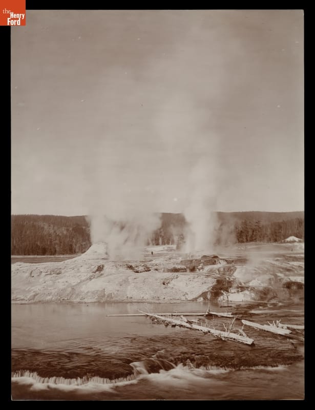Black-and-white photo of geysers erupting steam and/or water into the air in a rocky landscape with a lake or river in the foreground and forest in the background
