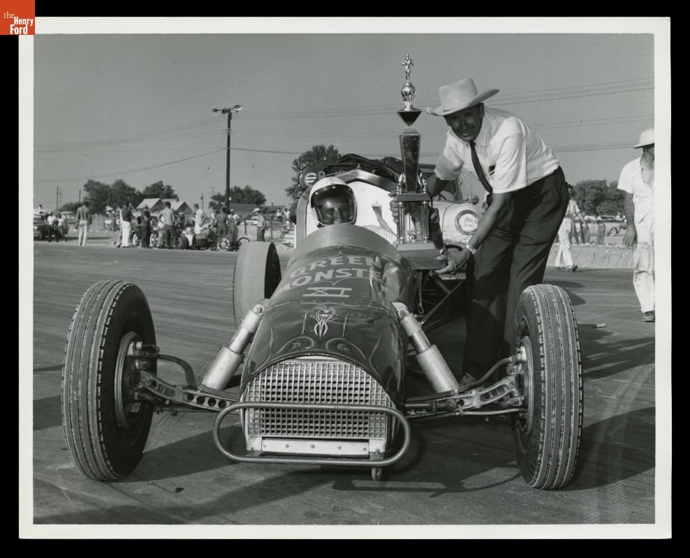 Man in cowboy hat bends down to hand large trophy to man in helmet in race car