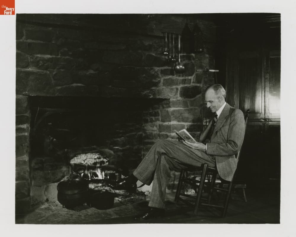 Henry Ford with a McGuffey Reader inside William Holmes McGuffey Birthplace, Greenfield Village, 1940 Man wearing suit sits in a chair by a fireplace, reading a book