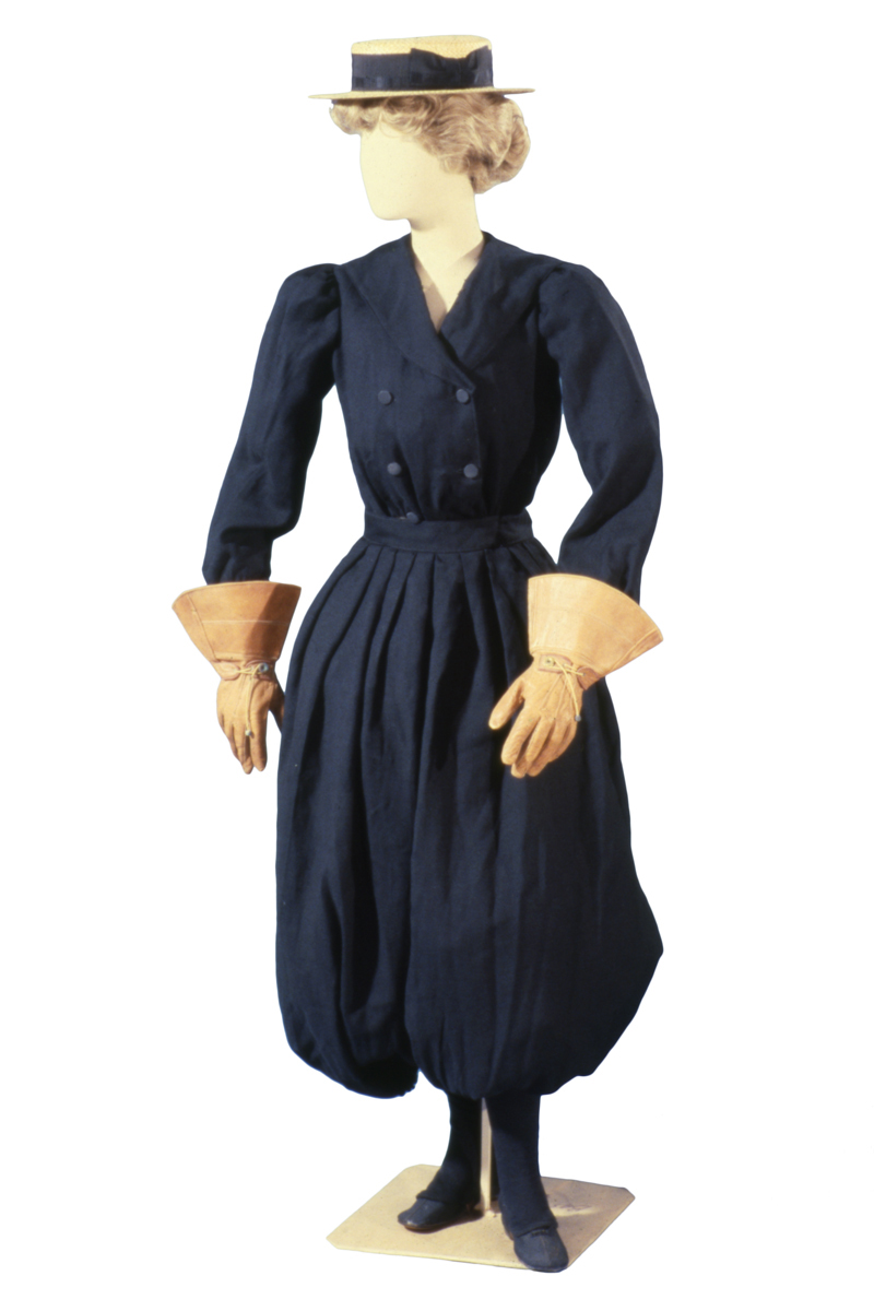 Mannequin wearing blue outfit with puffy bloomers, jacket with four button placket, large gloves, and a straw hat 