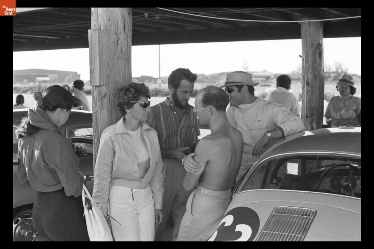 Group of people stand and talk under a pier or bridge surrounded by cars