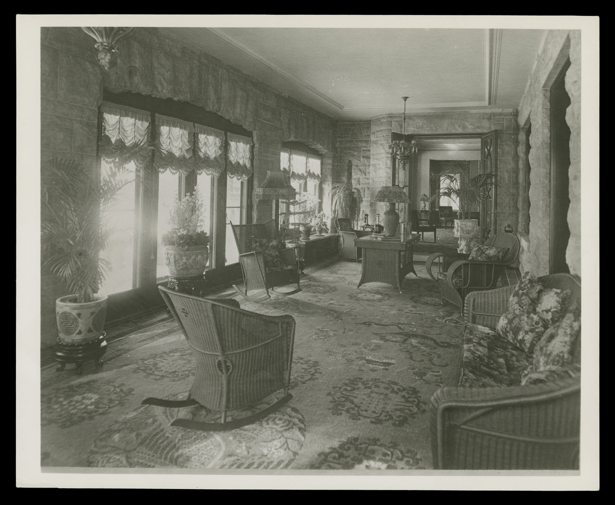 Black-and-white photo of long, narrow room filled with wicker furniture with many windows along one wall