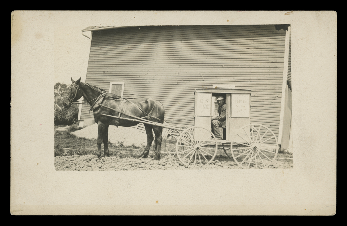 Man sits in very small boxy cart/wagon hitched to a horse in front of a building