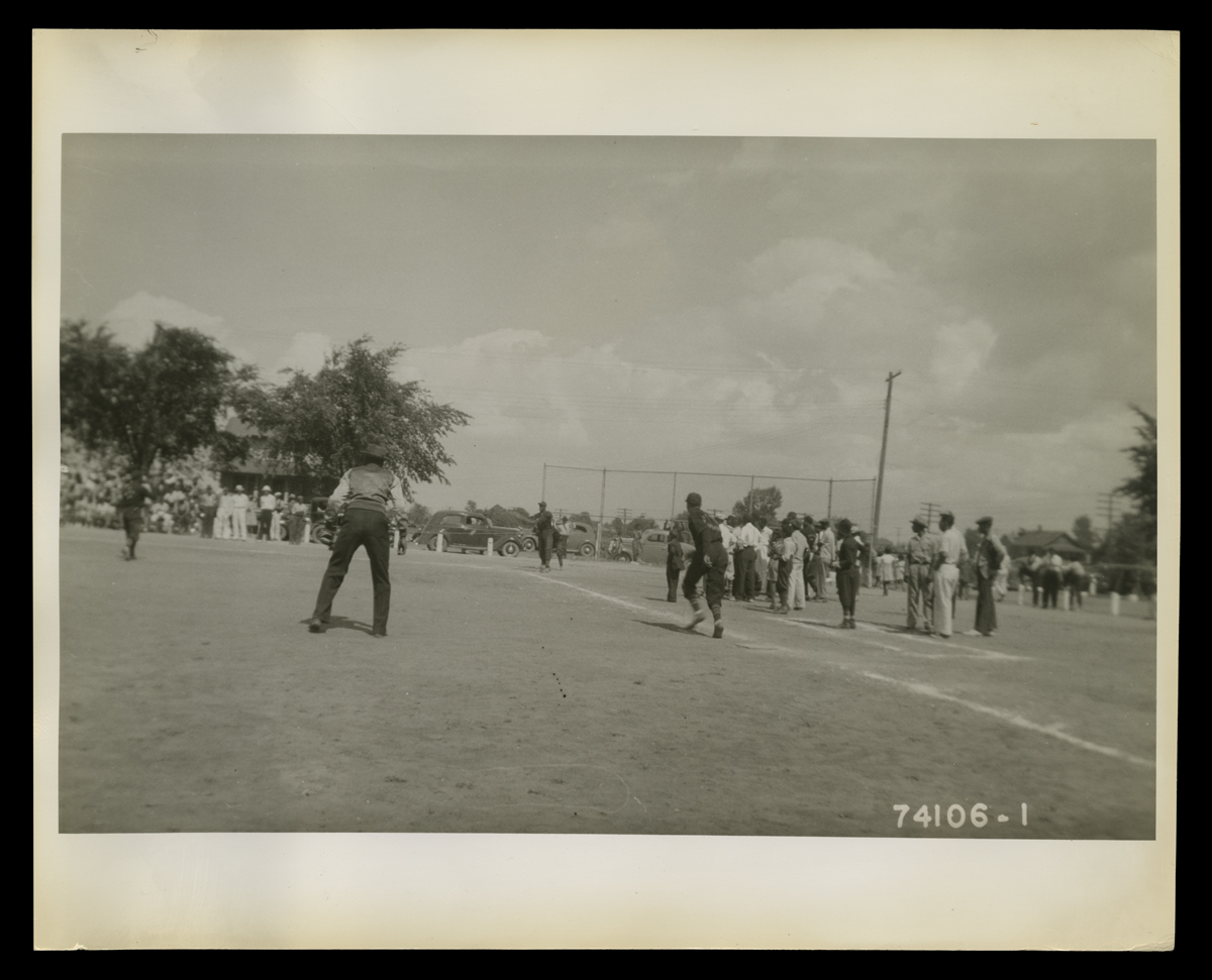 People, many or all African American, play baseball on a field while others look on