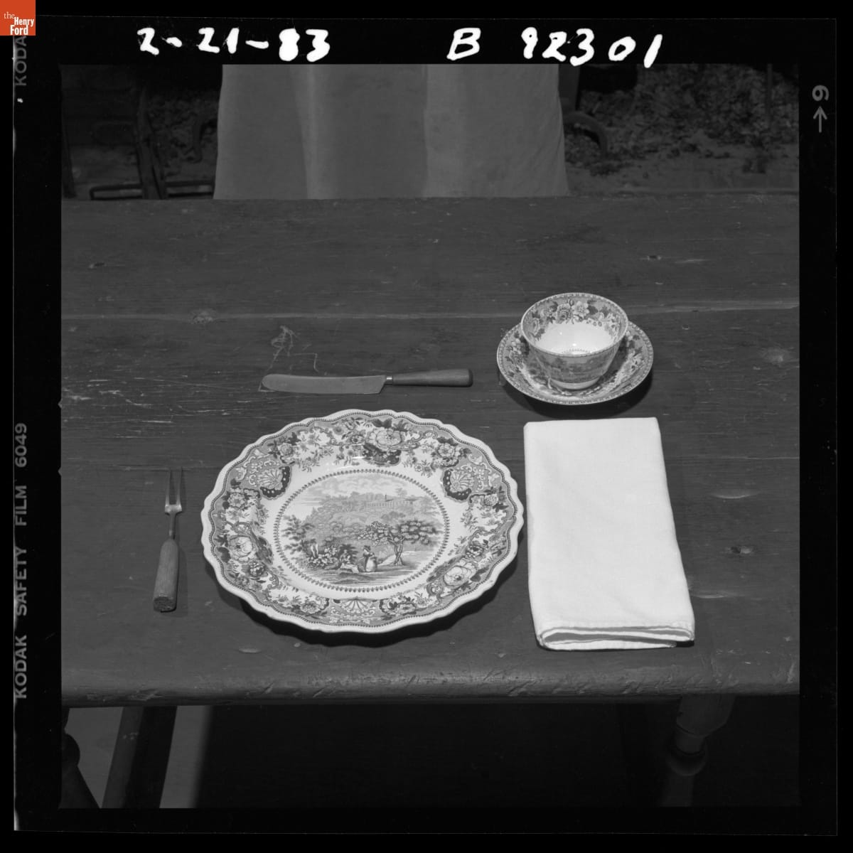 Table Setting in Eagle Tavern in Greenfield Village, February 1983 Black-and-white photo of table setting with plate, fork, knife, napkin, cup, and saucer on wooden table