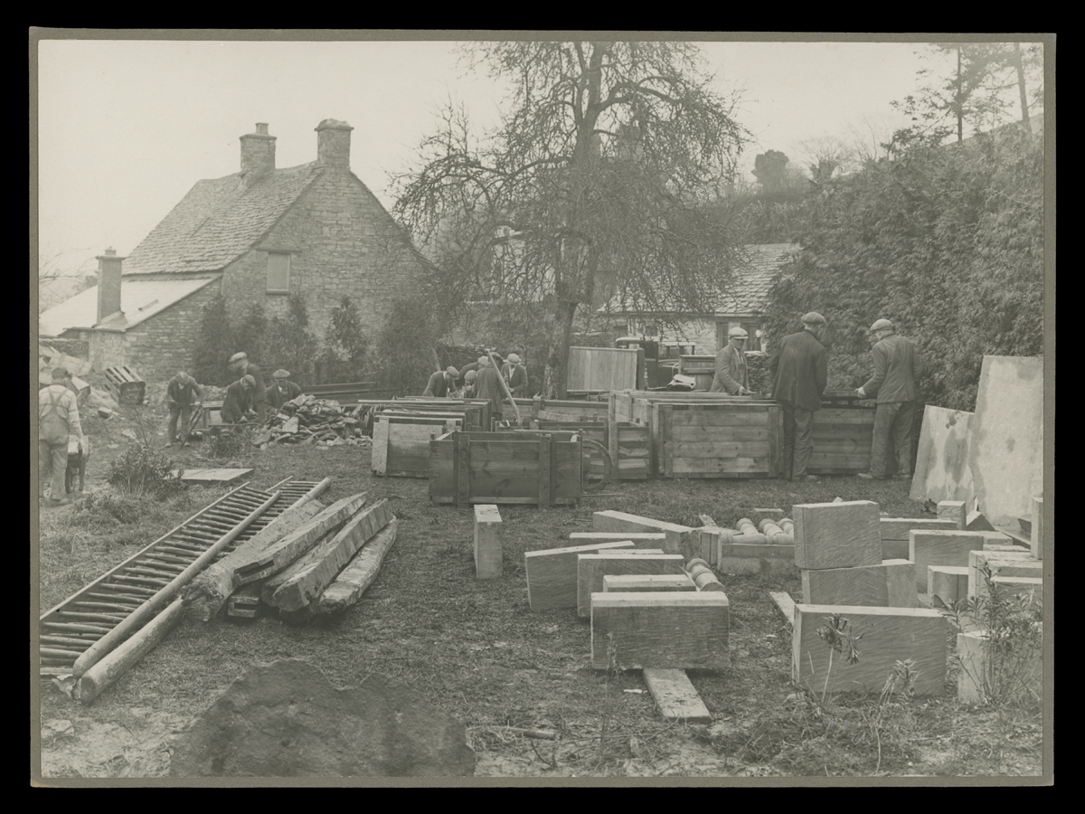 People around a group of crates and large stone building blocks in front of a stone house