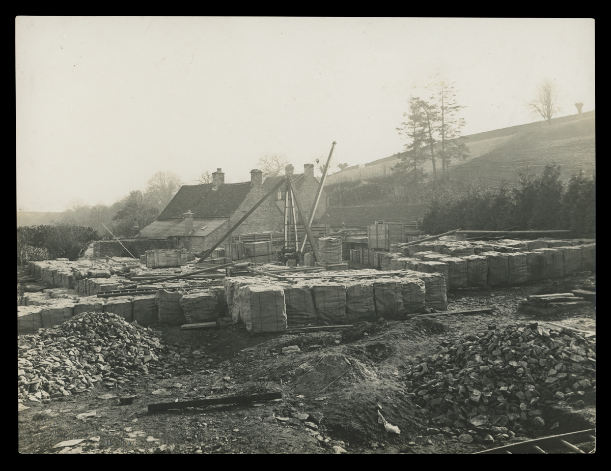 Construction site with piles of loose stones and many pallets; stone building in background