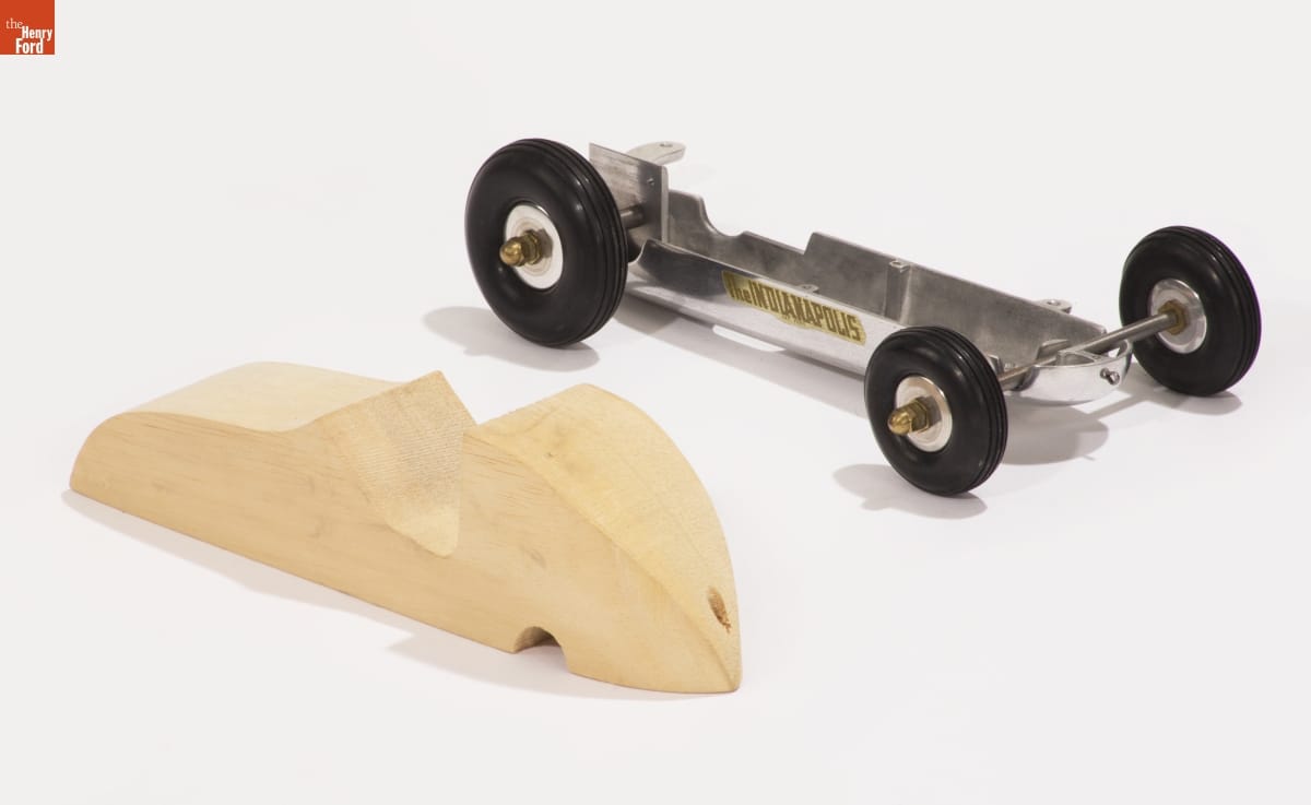 Korn Metal model car chassis sitting next to carved, unpainted block of wood in the shape of a car body
