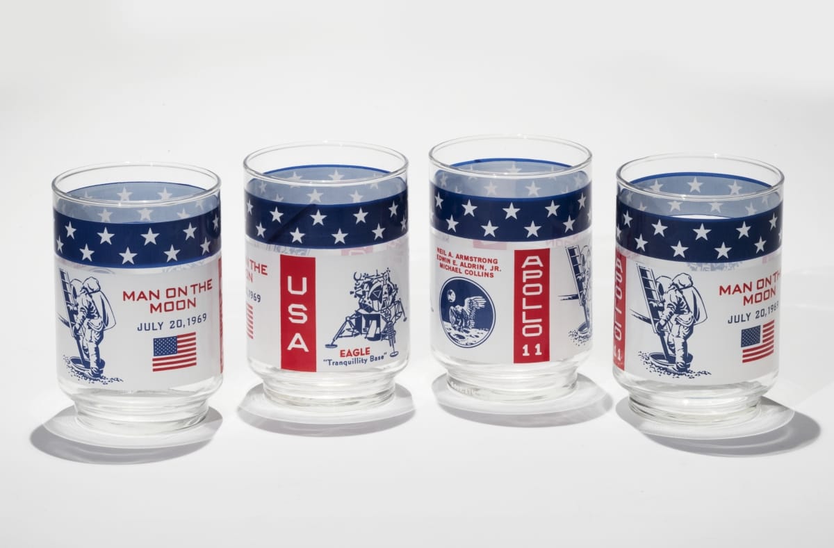 Four clear glasses with white, red, and blue designs, text, and spaceflight-related images