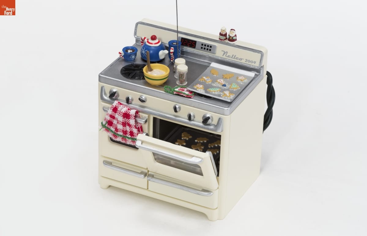 Ornament of cream-colored stove with one oven door cracked to show a tray of cookies inside; another tray of cookies, a bowl of dough, and a teapot and two mugs of hot chocolate are on top of stove